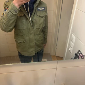Olivgrön jacka från Polo Ralph Lauren - Säljer en olivgrön jacka från Polo Ralph Lauren med coola patches på ärmarna och bröstet. Jackan har en hög krage och flera fickor framtill. Perfekt för en avslappnad och stilren look. Pris är inte hugget i stenen. Passar 