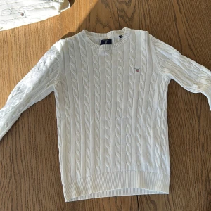 Vit stickad tröja från GANT - Säljer en stilren vit stickad tröja från GANT med klassiskt flätmönster. Tröjan har långa ärmar och en rund halsringning. Perfekt för sommarn 