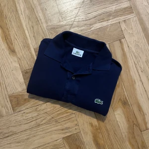 Mörkblå pikétröja från Lacoste - Säljer en klassisk mörkblå pikétröja från Lacoste med den ikoniska krokodilloggan på bröstet. Tröjan har korta ärmar och en stilren krage med knappar. Perfekt för en avslappnad men ändå stilren look.
