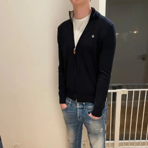 Morris Full zip - Säljer en mörkblå full zip från Morris med dragkedja och liten broderad logga på bröstet. Tröjan har lång ärm och är tillvärkaf i marino ull. Perfekt till jeans och t-shirt för en stilren look. Dock så har den ett litet hål på vänstra ärmen som man enkelt och smidigt kan sy ihop