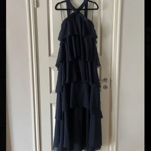 Mörkblå volangklänning från Vero Moda - Säljer en mörkblå långklänning från Vero Moda med flera lager volanger och korsade axelband. Perfekt för bal eller festliga tillfällen. Ny och oanvänd med lapp kvar! 