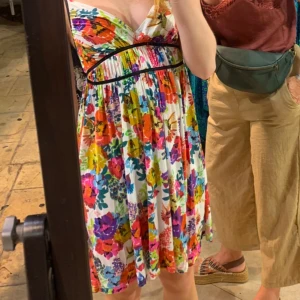 Blommig sommarklänning med v-ringning - Färgglad blommig klänning med v-ringad hals och smala axelband. Klänningen har en vit bas med stora blommor i rosa, lila, gul, grön och röd. Den är lätt utställd och slutar ovanför knäna, perfekt för varma dagar.