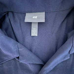 Mörkblå pikétröja från H&M - Säljer en stilren mörkblå pikétröja från H&M i storlek L. Tröjan har klassisk krage och knäppning med två knappar framtill. Perfekt för en clean och enkel look.
