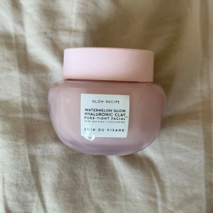 Watermelon Glow Hyaluronic Clay från Glow Recipe - En ansiktsmask från Glow Recipe med hyaluronsyra och vattenmelonextrakt. Denna lermask är designad för att förfina porerna och ge en jämnare hudton. Förpackningen är rosa och burken har en stilren design.