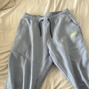 Ljusblå mjukisbyxor från Nike/tech - Säljer ett par ljusblå mjukisbyxor från Nike med elastisk midja och svarta snören. Byxorna har en gul Nike-logga på framsidan och svarta dragkedjor på fickorna. Perfekta för en avslappnad stil.