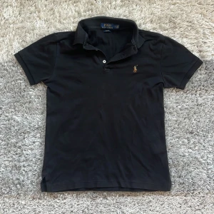 Svart pikétröja från Ralph Lauren - Säljer en stilren svart pikétröja från Ralph Lauren. Tröjan är slim fit och har en klassisk krage med knappar. Den är dekorerad med det ikoniska broderade polospelar-emblemet på bröstet. Perfekt för en avslappnad men ändå elegant look. Skriv gärna för fler frågor!