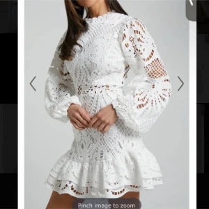 Vit spetsklänning med volang och lång ärm - Superfin vit kort klänning i spets med volangkant och ballongärmar. Hög hals och markerad midja, dragkedja bak. Perfekt för sommarens tillfällen eller student. Snyggt mönster och genombrutna detaljer. Superfin studentklänning!