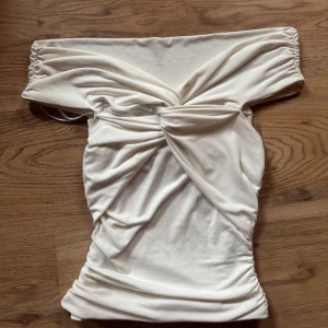 Vit offshoulder topp från BikBok - Säljer en vit offshoulder topp från BikBok i storlek XS. Tight passform med snygg rynkdetalj och twist framtill. Perfekt för dig som vill ha en stilren och trendig look.