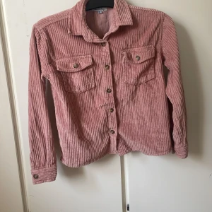 Rosa manchester-skjorta från Denim Day - Säljer en rosa manchester-skjorta från Denim Day med långa ärmar och två bröstfickor med knappar. Skjortan har klassisk krage och knäppning framtill. Perfekt för dig som gillar en avslappnad och trendig stil. 146-152