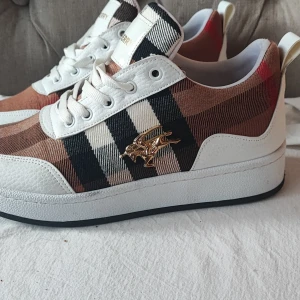 Rutig sneakers med vita detaljer från Burberry - Säljer ett par snygga sneakers från Burberry med klassiskt rutigt mönster i brunt, svart, vitt och rött. Skorna har vita snören, vit sula och guldfärgad logga på sidan. Perfekta för dig som vill sticka ut med stil.