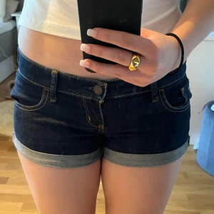 Mörkblå jeansshorts från Hollister - Säljer ett par mörkblå jeansshorts från Hollister med klassisk femficksdesign och uppvikta benslut. Shortsen har snygga kontrastsömmar och låg midja. Perfekta för varma dagar och enkel att matcha med olika toppar.