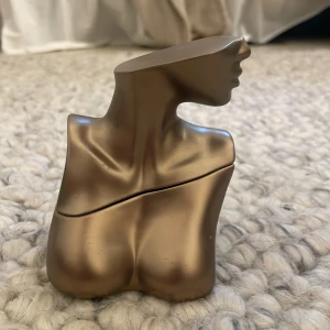 Billie Eilish parfymflaska i bronsfärgad form 50 ml - Unik parfymflaska från Billie Eilish, formad som en stiliserad kvinnobyst i bronsfärgad metall. Perfekt för dig som gillar vanilj och konstnärliga parfymer. Flaskan har konstnärlig design med mjuka linjer och detaljerat ansikte. 