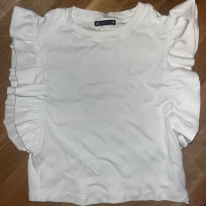 Vit topp med volangärmar från Zara - Säljer en vit topp från Zara med snygga volangdetaljer längs ärmarna. Toppen har rund halsringning och är tillverkad i ett mjukt, ribbat material. Perfekt för dig som gillar en feminin och trendig stil!