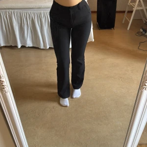 Svarta kostymbyxor - Snygga svarta kostymbyxor med hög midja och en klassisk straight passform. Perfekta för en stilren look. Byxorna har en enkel design med knappar och dragkedja framtill. Den korta modellen.