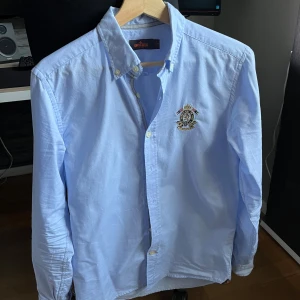 Ljusblå skjorta från Morris - Snygg ljusblå skjorta från Morris med broderad detalj på bröstet. Skjortan har långa ärmar och knappar framtill. Perfekt för en stilren look.