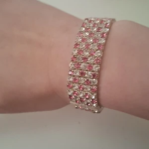 Glittrigt armband från SNÖ - Snyggt glittrigt armband från SNÖ med rosa och klara stenar. Armbandet har en bred design och är perfekt för att ge en extra touch till din outfit. Det elastiska bandet gör det enkelt att ta på och av.
