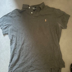 Grå pikétröja från Polo Ralph Lauren - Säljer en klassisk grå pikétröja från Polo Ralph Lauren med korta ärmar och en broderad logotyp på bröstet. Perfekt för en stilren och avslappnad look.