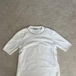 Säljer en stilren vit tröja från COS med korta ärmar och en subtil svart kant runt halsen. Perfekt för en minimalistisk look. Tröjan är i ett mjukt material som ger en bekväm passform.