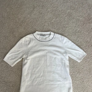 Vit kortärmad tröja från COS - Säljer en stilren vit tröja från COS med korta ärmar och en subtil svart kant runt halsen. Perfekt för en minimalistisk look. Tröjan är i ett mjukt material som ger en bekväm passform.