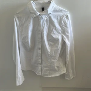 Vit skjorta från H&M - Klassisk vit skjorta från H&M med långa ärmar och knappar framtill. Perfekt för en stilren look. Skjortan är tillverkad i bomull och har en traditionell krage. Finns lite små bruna prickar på framsidan under bysten och på baksidan av ena armen men bör inte märkas särskilt mycket i vardagen. Dessutom kan de förmodligen försvinna vid blekning eller liknande. Lite mindre i passform så kan fungera på mindre storlekar