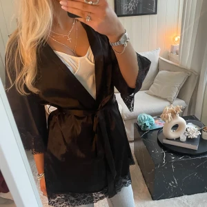 Svart kimono med spetsdetaljer - Elegant svart kimono med knytband i midjan och vackra spetsdetaljer vid ärmslut och nederkant. Perfekt för en stilfull look.