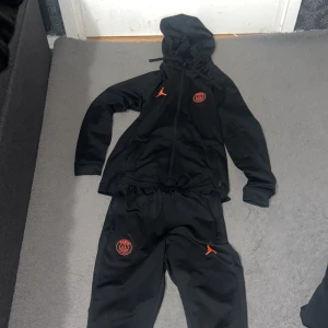 Psg tracksuit  - Säljer ett svart träningsset från Jordan i samarbete med PSG. Setet består av en jacka med dragkedja och byxor, båda med PSG-logga och Jordan-märke i rött. Jackan har huva och byxorna har elastisk midja med dragsko.