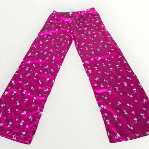 Rosa byxor med Snoopy-mönster - Säljer ett par rosa byxor med ett gulligt Snoopy-mönster. Byxorna har en loose passform och är tillverkade i ett glansigt material. Perfekta för en avslappnad stil med en rolig touch! Storlek 16, tror det motsvarar M.