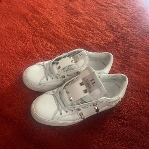 Vita Valentino Rockstud sneakers - Säljer ett par vita Valentino Rockstud sneakers med ikoniska nitar i silver längs sidorna och på plösen. Skorna har snörning och är tillverkade i skinn med rund tå och platt sula. Perfekta nu till våren och sommaren. Priset är inte hugget i sten 