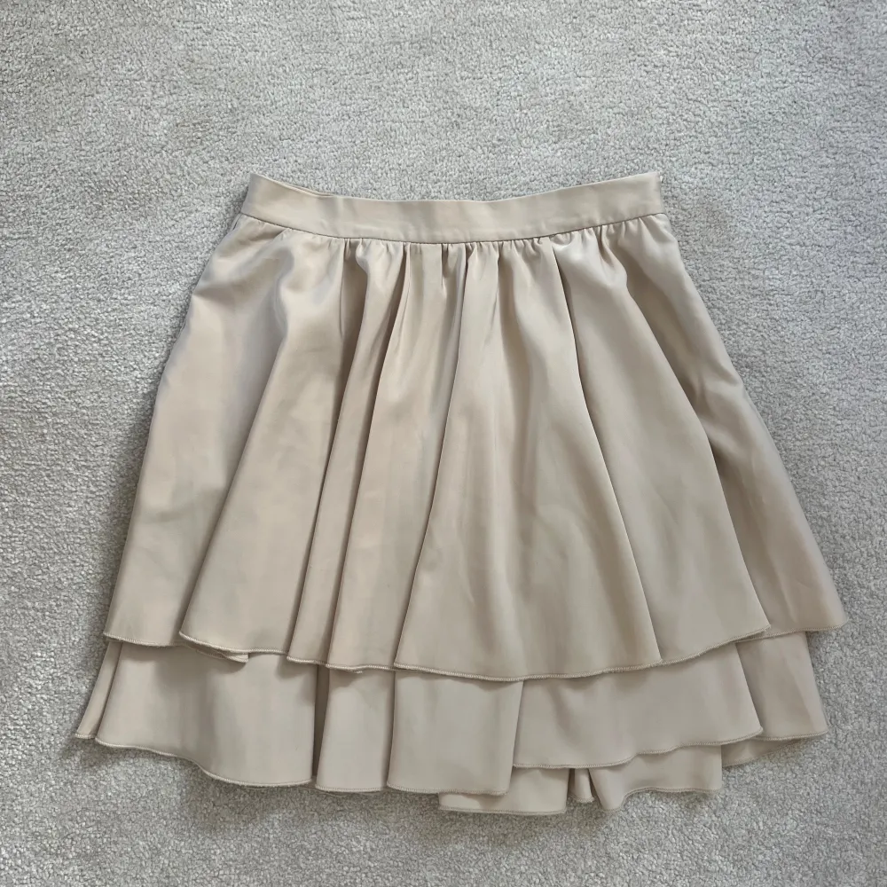 Säljer en superfin beige volangkjol från NAKD. Kjolen har flera lager och ett snyggt fall, perfekt för dig som gillar en feminin och trendig stil. Mjuk i materialet, midjelinningen är enkel och bekväm. Nån svag fläck, ej försökt få bort i tvätten . Hameet.