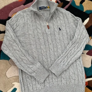 Grå stickad polotröja från Polo Ralph Lauren - Säljer en klassisk grå stickad polotröja från Polo Ralph Lauren med kabelstickat mönster och halv dragkedja. Tröjan har lång ärm, ribbade muddar och den ikoniska broderade loggan på bröstet.användt 3 gånger skriv pm för mer info