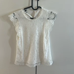 Vit spetsblus från PIECES - Säljer en söt vit blus från PIECES i spets med fina detaljer och korta ärmar. Blusen har knapp i nacken och vackert mönstrad spets längs kanterna. Perfekt för dig som gillar romantisk stil!