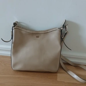 Beige axelväska från Coccinelle - Stilren beige axelväska från Michael Kors med justerbar axelrem och diskret logga framtill. Väskan har dragkedja upptill och är tillverkad i slätt skinn. Perfekt för dig som vill ha en klassisk och tidlös accessoar.