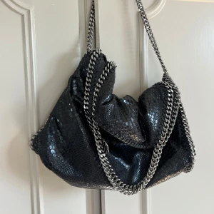 Svart axelväska från Stella McCartney - Snygg svart axelväska från Stella McCartney med ett ormskinnsmönster. Väskan har vissa slitningar (se bild 4 och 5). Väljer man att vika ner väskan (se bild 1) så syns inte slitningarna på bild 5! Väskan är rymlig och har en innerficka! 
