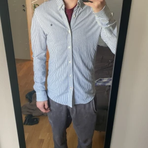 Vit och blå randig skjorta  - Klassisk vit och blå randig skjorta från Morris med knappar hela vägen och broderad logga på bröstet. Skjortan har lång ärm och normal passform, perfekt till många olika tillfällen. Den är i storlek M och i väldigt bra skick. Hör av er vid funderingar