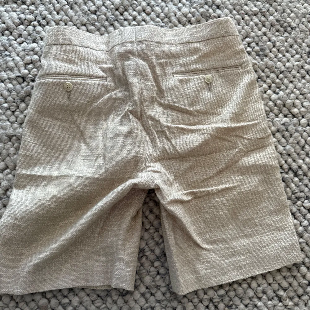 Stilrena beiga shorts i linnematerial. Klassisk passform med veck framtill, knapp och dragkedja i midjan samt två bakfickor med knappar. Perfekta för varma dagar.. Shortsit.