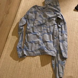 Underställströja och byxor från Cross Sportswear - Säljer en blågrå långärmad underställströja och byxor från Cross Sportswear med camouflagemönster. Tröjan är tillverkad i en mjuk blandning av polyester, nylon och elastan, perfekt för kalla dagar och aktiviteter utomhus.