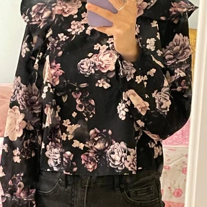 Svart blommig blus med volang från KappAhl - Supersnygg svart blus från KappAhl med lila och rosa blommor. Blusen har långa ärmar och volangdetaljer vid axlarna och framtill. Perfekt för dig som vill sticka ut lite extra med ett romantiskt mönster!