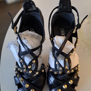 Svarta klackskor med nitar från Eden - Säljer ett par svarta klackskor sandaletter från Eden med smala remmar och guldfärgade nitar. Skorna har öppen tå och hög klack, tillverkade i äkta skinn. Extra yttersula påsatt. Snyggt med spänne vid ankeln.