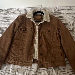 Brun manchesterjacka med teddyfoder från 157 - Säljer en brun manchesterjacka från 157 med vitt teddyfoder och krage. Jackan har knappar framtill, två bröstfickor med lock och klassisk jeansjacke-look. Perfekt för kyligare dagar och ger en cool vintagekänsla.