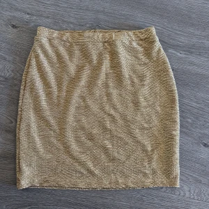 Guldig glittrig kjol från H&M - Supersnygg guldig kjol från H&M med glittrigt tyg och rak passform. Perfekt för dig som vill sticka ut lite extra. Kjolen är kort och har en elastisk midja för skön passform.