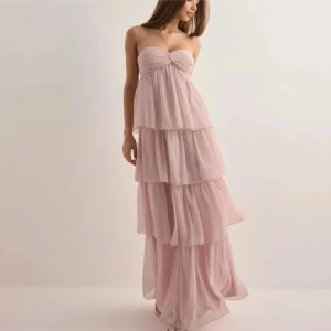 Ljusrosa axelbandslös långklänning - Elegant ljusrosa långklänning med volanger och axelbandslös design. Perfekt för speciella tillfällen med sin flytande och romantiska stil. Balklänning från nelly. Ny med prislapp!