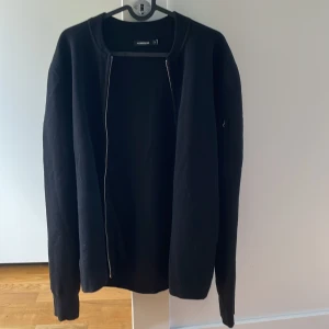 J.Lindberg Kofta (andon black zip cardigan) - En helt oanvänd J.Lindeberg kofta i svart, medium storlek. Denna andon zip cardigan är en stilren och tidlös tröja som passar perfekt för alla tillfällen. Den har aldrig blivit använd och är i utmärkt skick. 