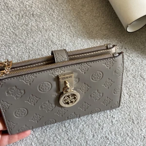 Guess plånbok/väska - Guess plånbok/väska i en lung grå färg, med guess mönster över hela plånboken. Den är såklart äkta och köpt via Zalando. Qr kod finns. Använt 3-4 gånger. På ena sidan ser man lite slitning/repor på skinnet. Annars väldigt fräsch och fin. Nytt pris 899kr.