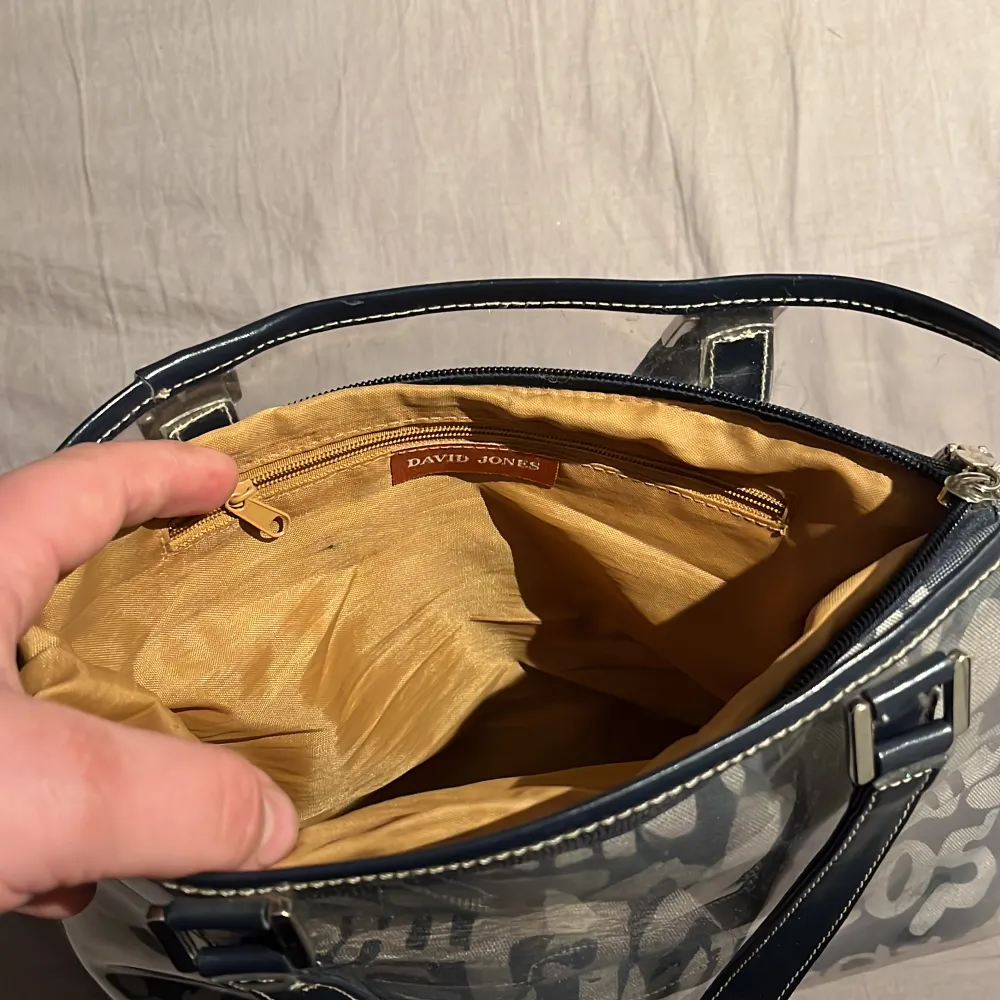 En två delad handväska genomskinlig gummi handväska med passande necessär som går att ta ut, handväskan har mörkblåa detaljer och den stängs med dragkedja . Väskor.