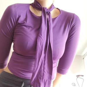 Y2K topp med halsband - Inköpt mellan år 2000-2010. Så unik och fin Y2K topp i en härligt lila färg och med ett band som sitter fast och knyts runt halsen. Ganska glatt tygg som är supermjukt. Har en söm som gått upp på ena armslutet, går lätt att sy (själv saknar jag tråd i rätt färg). Svarar på alla frågor.  
