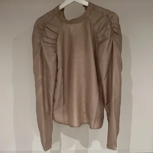 Beige glänsande blus - Blus från Vila som är beige med lite metallic lyster. Lång ärm med lite ”rynkor” vid axeln. Knäpps med två knappar i nacken till en liten ståkrage. Fin till vardags och fest. 