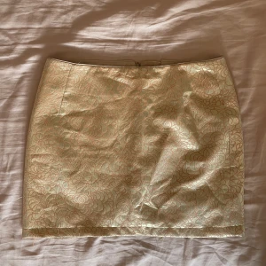 Kort guldig kjol med blommönster - Kort kjol i guld med rosa/beige blommigt mönster. Den är uppsydd lite av mig. Jag är 165 cm och brukar ha storlek s-m. Kan mötas nära Mariatorget. 