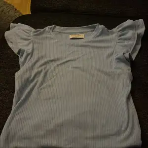 Säljer denna ljusblåa t-shirten med volanger på ärmarna. Den kommer inte till användning längre och har bara använt den 1 gång. Väldigt bra skick. Storlek Small