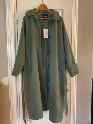 Grön kappa från Monki - Helt ny, lång kappa med prislappen kvar. Storlek L. Jättefin grågrön färg, men tyvärr inte en modell som passar mig. 