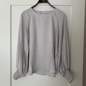 Blus, Ellos, Beige/creme, stl 36 - Blus i vävd kvalitet med lyster. Rund hals, sprund och knapp i nacken. Långa ballongärmar med smala ärmslut i smock. Längd ca 62 cm i stl 36. Använd 1 gång.